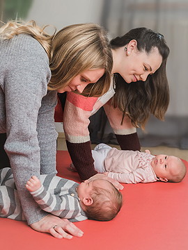 zwei Babys auf dem Rücken liegend am Boden, Mütter auf Tuchfühlung knieend über ihnen.
